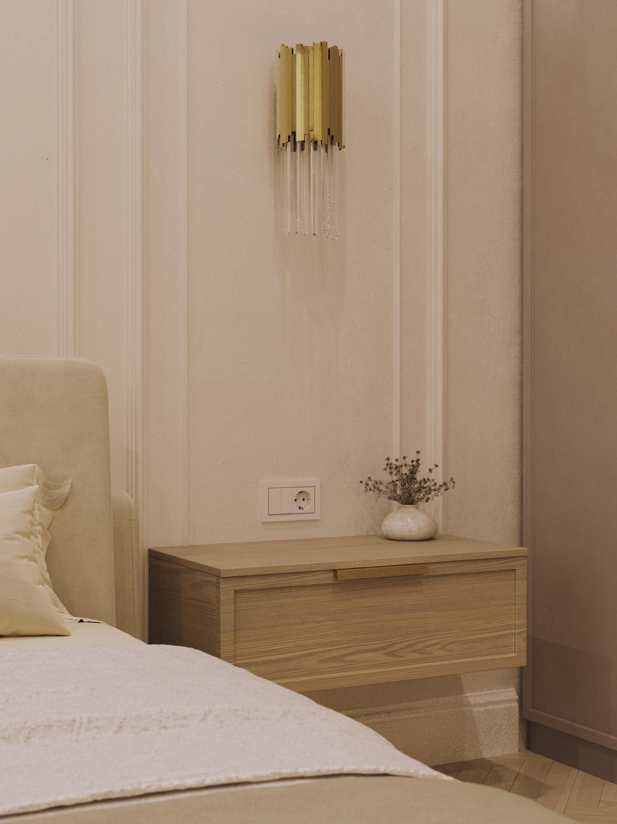 Modern bedroom with light wooden nightstand, gold wall sconce, and white bedding.